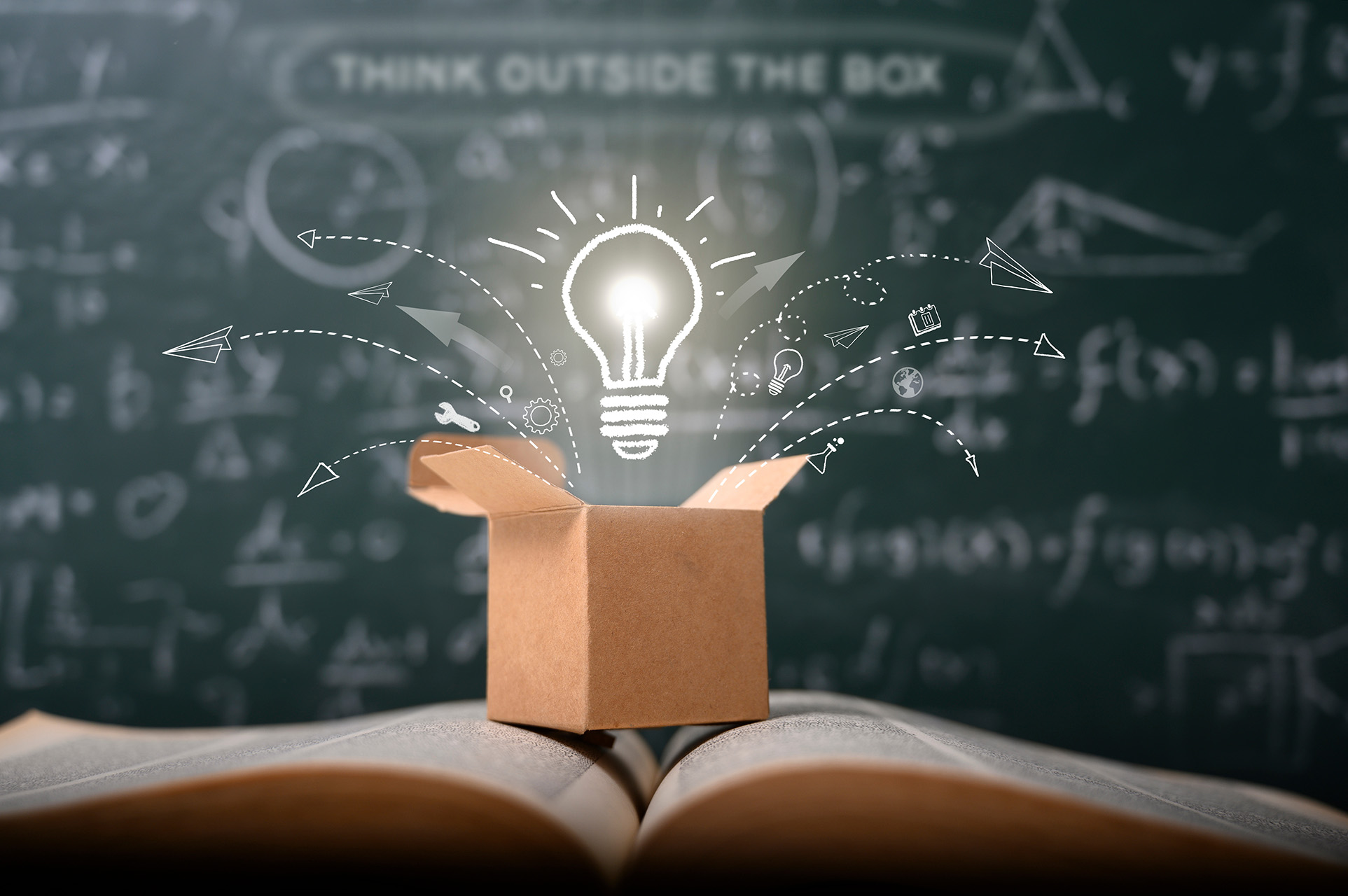 Innovate Core Values A small cardboard box with its top flaps open sits on top of an open book featuring an illustration of an emerging, glowing light bulb, which represents one of Emerald's core values: innovation.