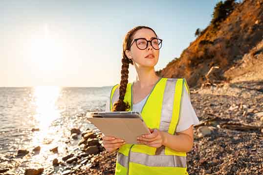 Environmental and Brand Protection Benefits of Product Destruction Woman standing by the lake shore in a white shirt and yellow safety vest, holding an iPad to represent environmental and brand protection